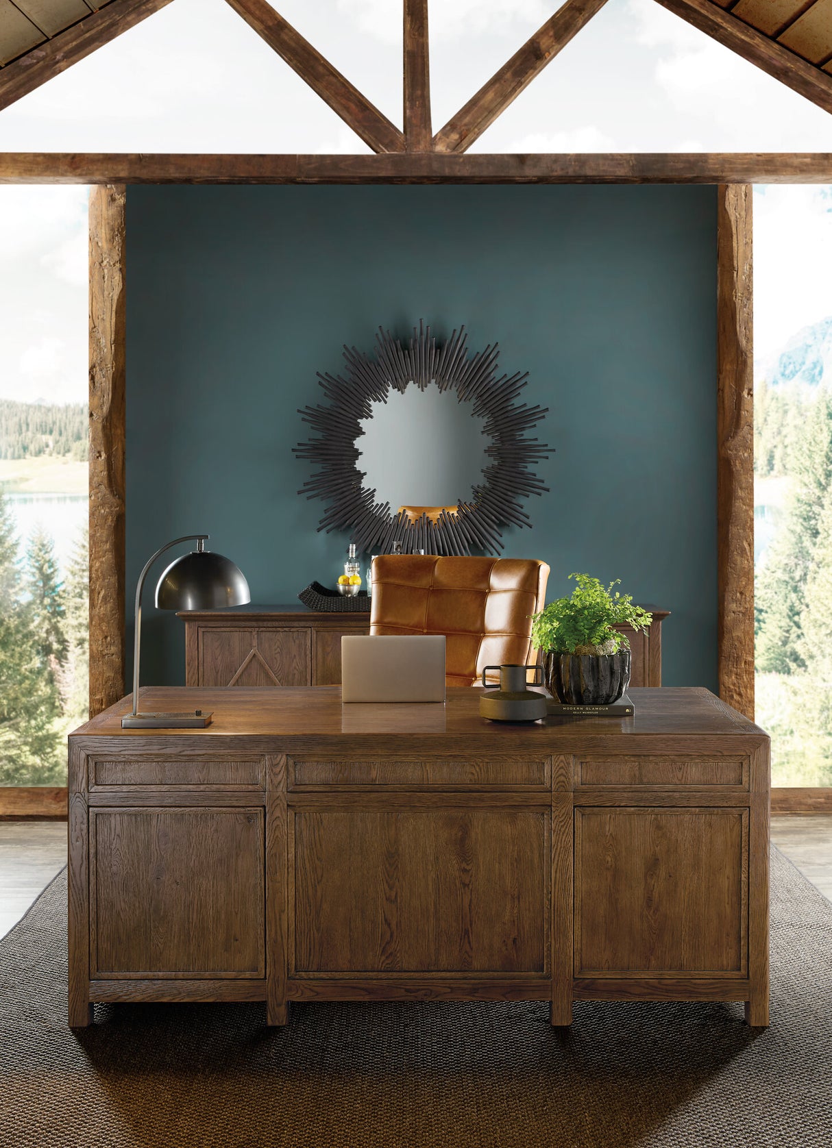 Wooden desk with a laptop, mouse, and decorative items in a room with large windows and a decorative mirror on the wall.