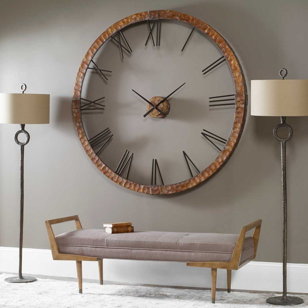 Large wall clock with Roman numerals in a room with a bench and lamps.